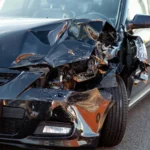 Compra de auto chocado: Primer plano de un vehículo oscuro con el capó y el parachoques aplastados tras un impacto frontal, mostrando daños extensos.