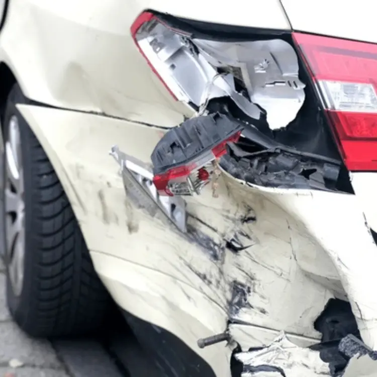 Compra de auto chocado: Primer plano de la parte trasera de un coche blanco con el parachoques y la luz trasera severamente dañados, indicando un impacto posterior.